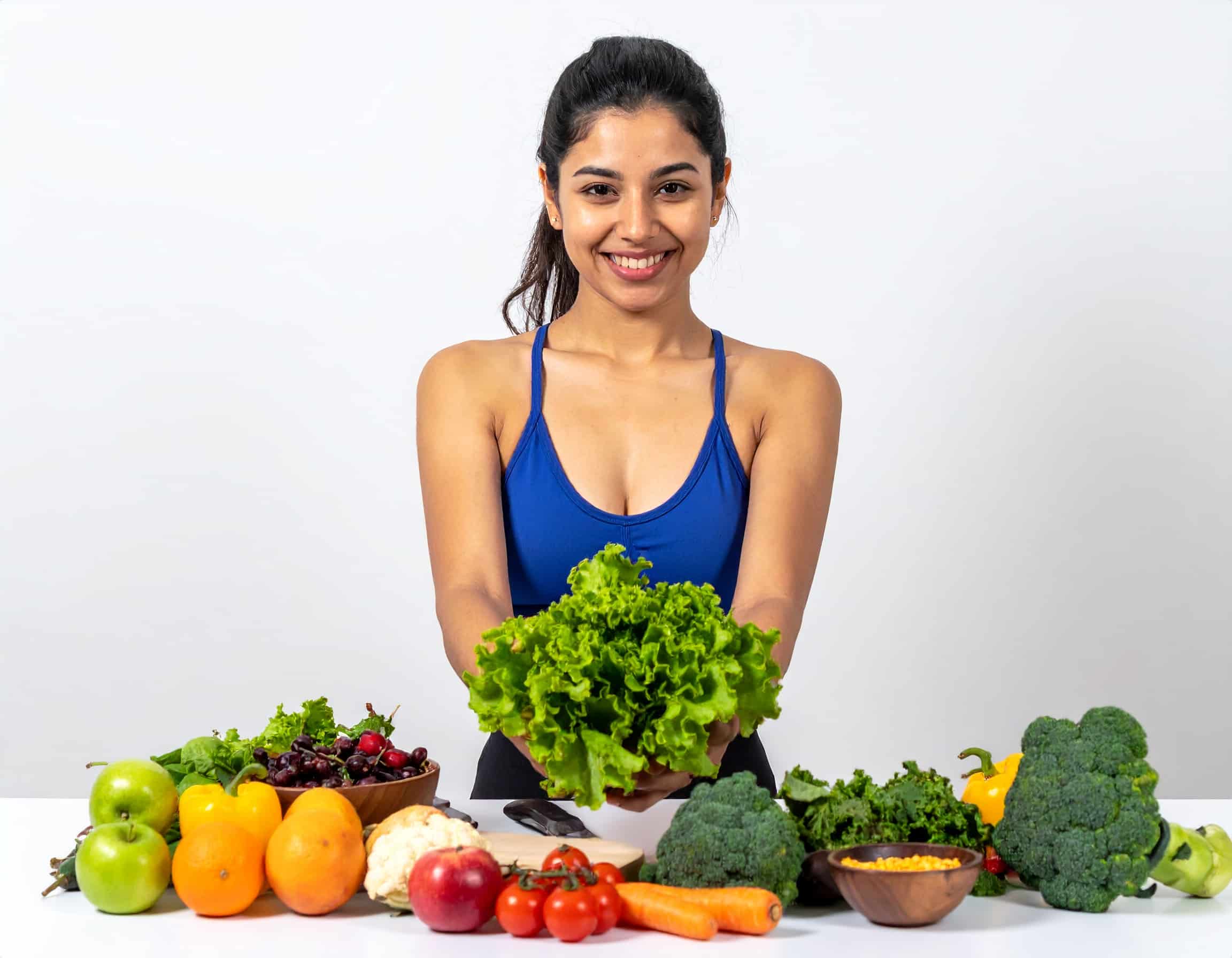 Eine lächelnde, sportliche junge Frau hält einen frischen grünen Salatkopf in den Händen. Vor ihr auf dem Tisch liegt eine bunte, vielfältige Auswahl an frischem Obst und Gemüse