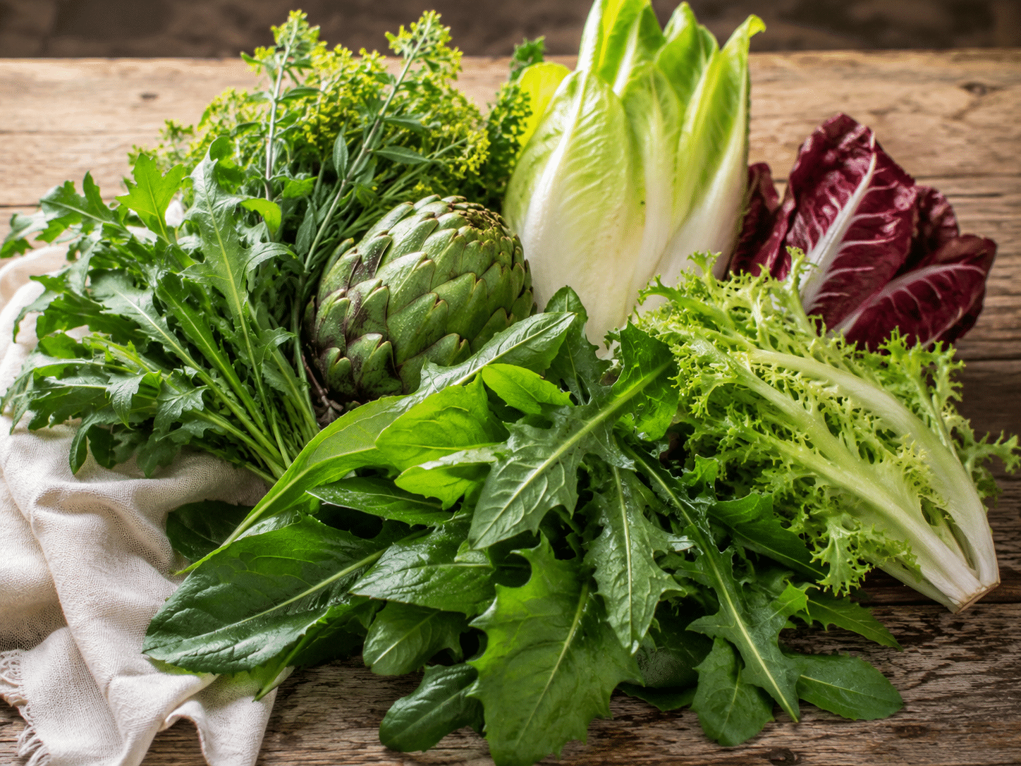 Eine rustikale Anordnung von bitterstoffreichen Gemüsesorten und Kräutern, darunter eine Artischocke, Radicchio, Chicorée, Endivie und Löwenzahnblätter auf einem Holztisch.