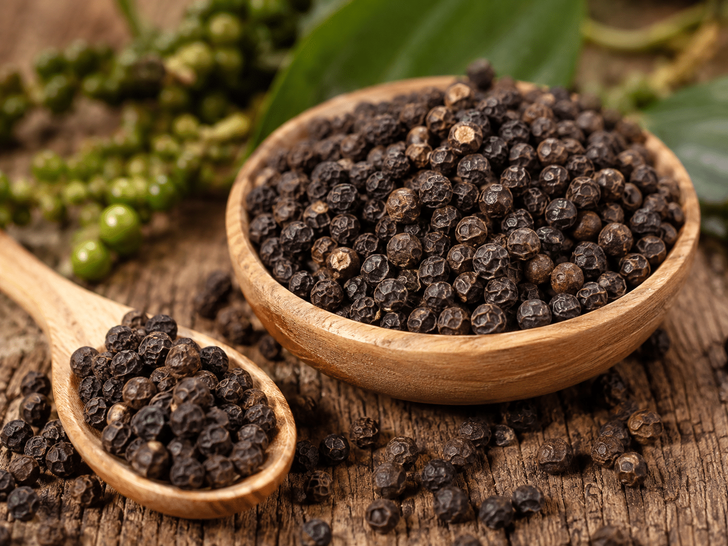 Getrocknete, schwarze Pfefferkörner in einer Holzschale und auf einem Holzlöffel. Im Hintergrund liegen frische, unreife grüne Pfefferbeeren an einer Rispe.