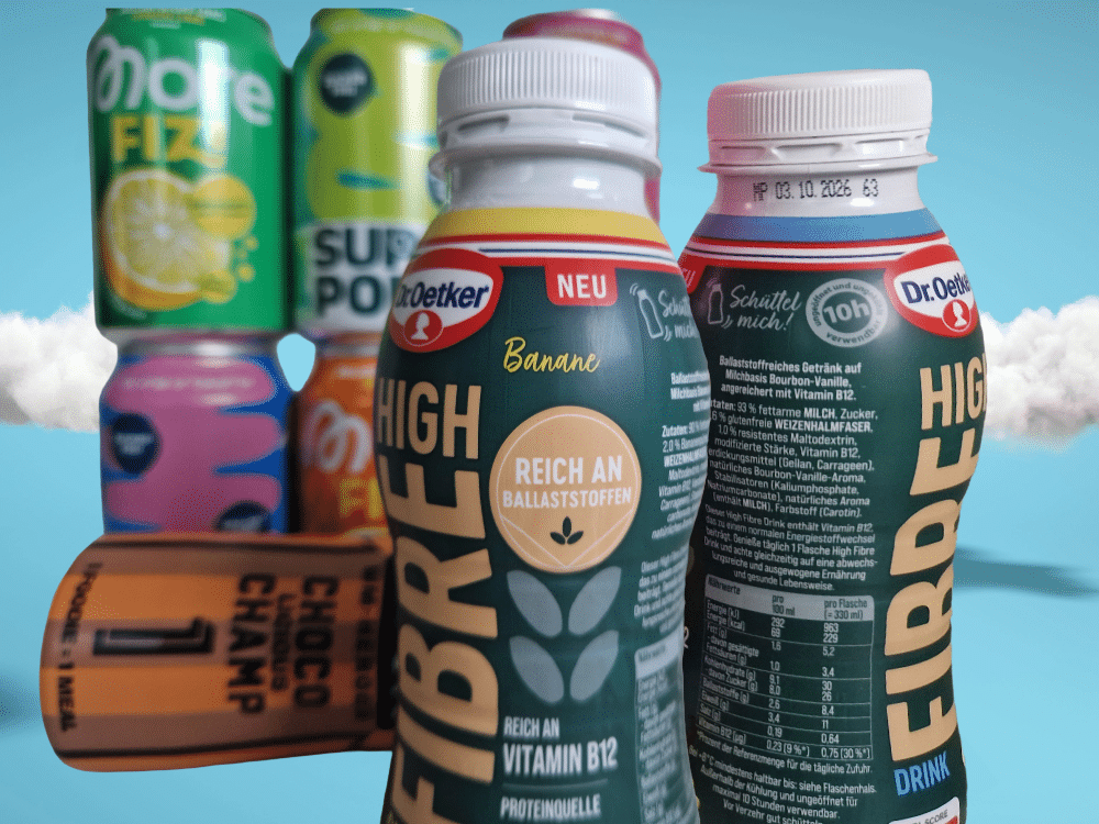 Zusammenstellung verschiedener Funktionsgetränke vor blauem Himmel: Im Vordergrund zwei Dr. Oetker High Fibre Flaschen und eine liegende Ehrmann Foodie Flasche, dahinter gestapelte Dosen von More FIZI und Super Pop.