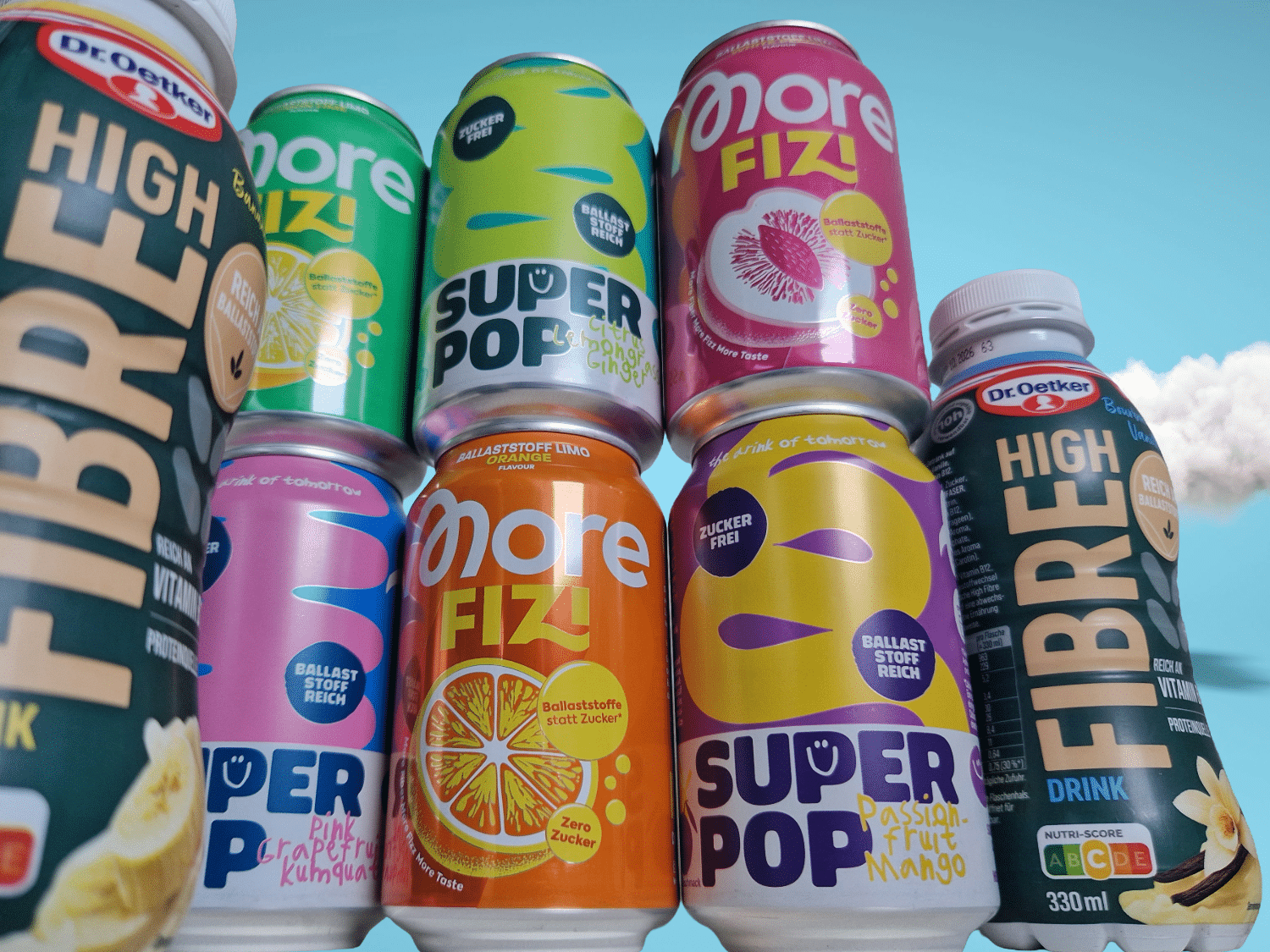 Ballaststoffdrinks im Check: Eine bunte Ansammlung von gestapelten Super Pop und More FIZI Dosen, flankiert von zwei Dr. Oetker High Fibre Flaschen vor blauem Himmel.